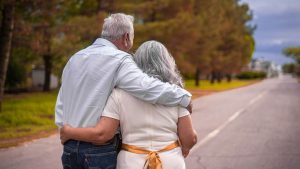 An older man and woman with grey hair walking arm in arm down a road