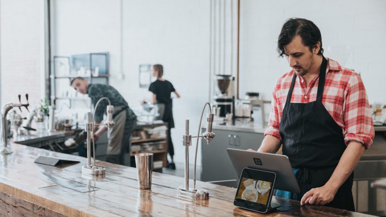 A barista in a plaid shirt and apron using a tablet at a wooden counter